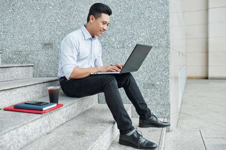Positive young Asian software developer sitting on steps and working on laptopの写真素材