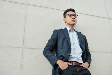 Confident young Vietnamese businessman in glasses putting hands in pockets when standing at building wall and looking in front of himの写真素材