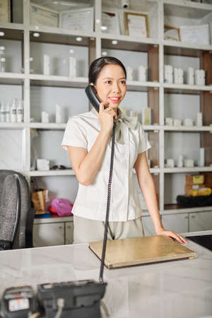 Smiling pretty spa salon worker standing at table and talking on phone with clientの写真素材