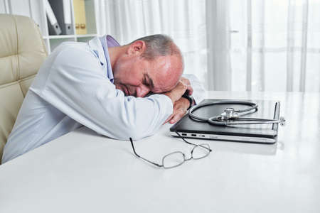 Mature general practitioner resting on his desk with laptop, stethoscope and glasses in front of himの写真素材