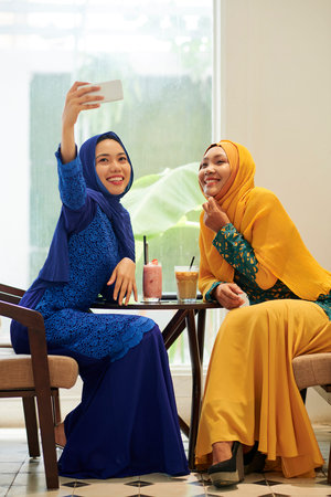 Happy young Asian muslim woman taking selfie with her friend when spending time in cafe
