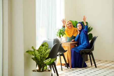 Smiling young muslim women in traditional dresses sitting at cafe table and waving with hands when trying to bring attention on waiterの写真素材