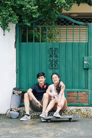 Smiling hugging teenage brother and sister sitting in front of house gates and resting after...