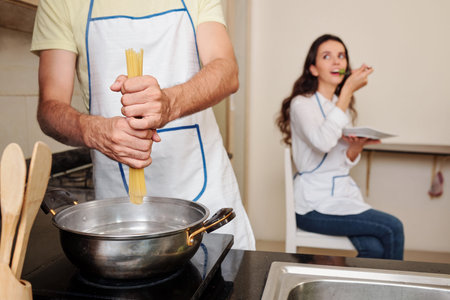 Young man putting spaghetti in boiling water when his wife eating salad in background