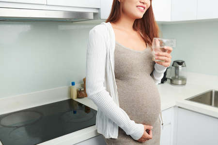 Cropped image of young pregnant woman touching her belly when drinking a glass of fresh water in kitchenの写真素材