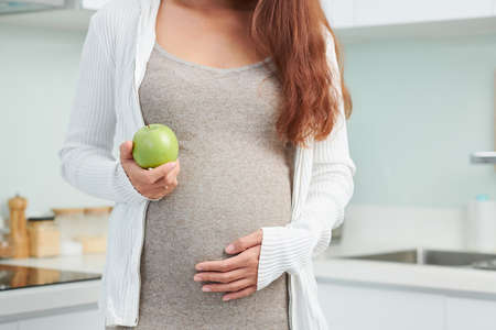 Cropped image of pregnant woman touching her big belly and showing fresh green apple she is having for snackの写真素材
