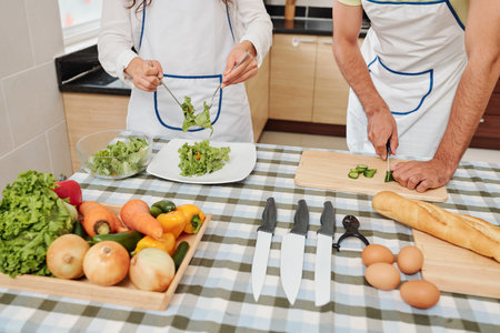 Couple cutting cucumbers and mixing lettuce leaves with dressing for delicious healthy salad for...