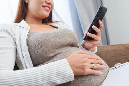 Close-up image of positive young woman touching her pregnant belly and checking social media on her smartphoneの写真素材