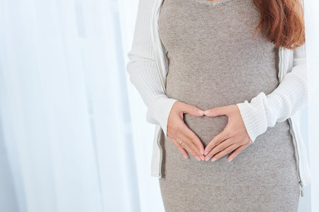 Pregnant woman in woll dress putting hands on her body and making heart shape with fingers