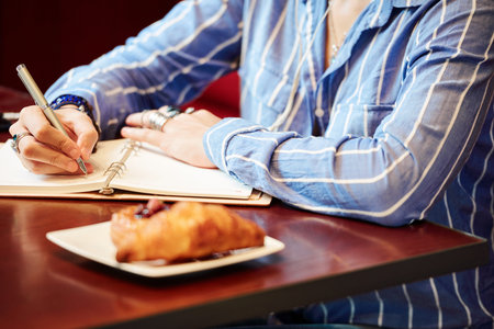 Hands of female student sitting at cafe table with pastry on plate and taking notes in textbookの写真素材