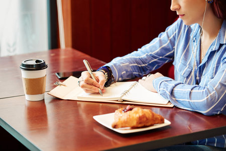 Cropped image of creative young woman sitting at table with coffee and croissant for breakfast and writing down her plansの写真素材