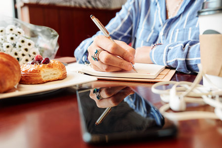 Close-up image of creative woman eating delicious pastries and writing plans and ideas in diary