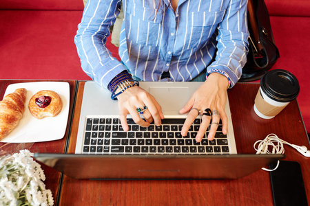Hands of creative young woman with many rings and bracelets sitting at table with coffee and...