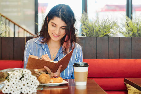 Pretty smiling young woman sitting at table in cafe with coffee and couquet of flowers and taking notes in diaryの写真素材