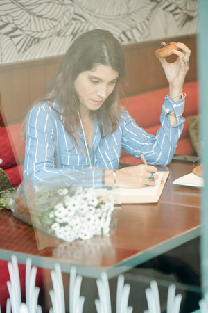 Creative young woman sitting at cafe table, eating cherry puff and writing thoughts in planner