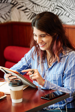 Laughing young woman sitting at cafe table and looking at funny mems on her tablet computerの写真素材
