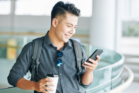 Positive young Asian man drinking take away coffee and reading text messages in his smartphone