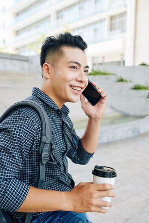 Attractive young Vietnamese man standing outdoors, talking on phone and drinking tasty take away...