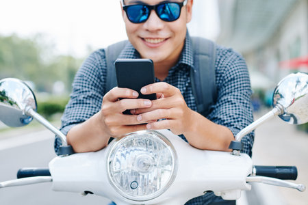 Happy young Asian man in sunglasses sitting on scooter and checking text messages and notifications...
