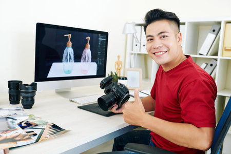 Portrait of happy young Asian photographer sitting at desk with camera in hands and showing...