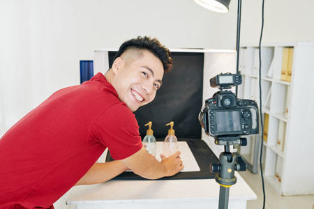 Happy young Asian man putting glass soap dispensers against black background to take photos for...
