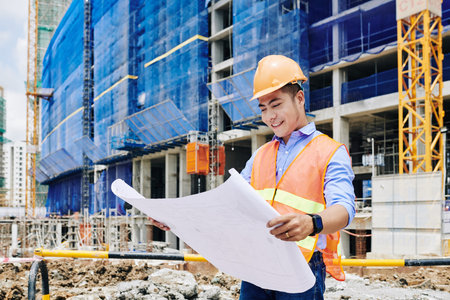 Smiling Asian construction engineer in orange vest and hardhat examining building blueprintの写真素材
