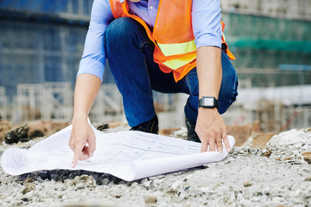 Cropped image of costruction worker pointing at bulding blueprint he spreaded out on the ground