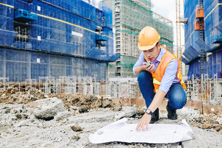 Professional Vietnamese engineer examining construction plan and talking on walkie-talkie with...