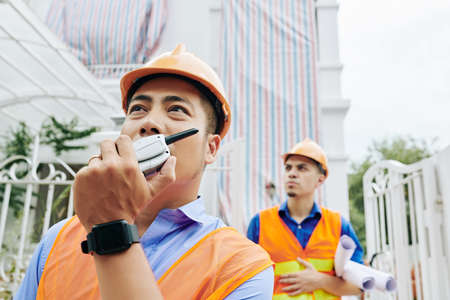 Serious Asian contractor controlling work at construction site by talking on walkie-talkie with colleaguesの写真素材