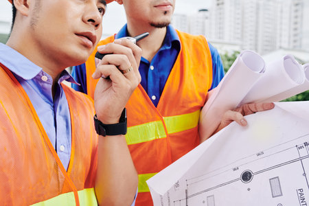 Cropped image of serious construction workers with building blueprint in hands talking on...