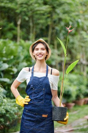 Portrait of young cheerful proud gardener in denim apron and gloves posing with flower pot outdoorsの写真素材