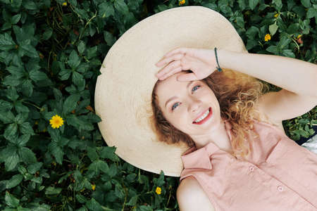 Happy young Caucasian woman in big straw hat lying on grass in park and lookig at camera, view from aboveの写真素材