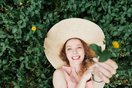 Happy pretty young Caucasian woman in big straw hat lying in grass and giving you beautiful flowerの写真素材