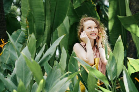 Happy young woman standing in tropicsl garden and listening to music in earbudsの写真素材