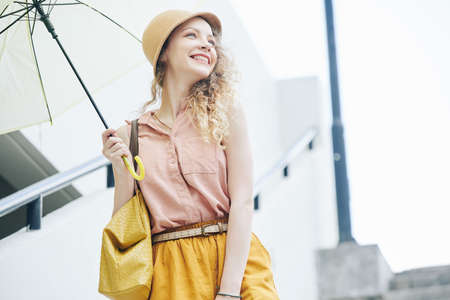 Attractive happy young woman in hat walking outdoors with umbrella on rainy dayの写真素材