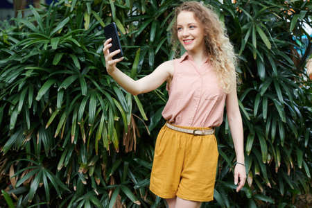 Lovely young smiling woman with blnd curly hair taking selfie againsts green busches in parkの写真素材