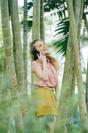 Happy young woman leaning on bamboo tree and talking on phone with friendの写真素材