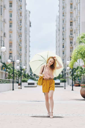 Happy pretty smiling girl in shorts and blouse walking in the city with umbrellaの写真素材