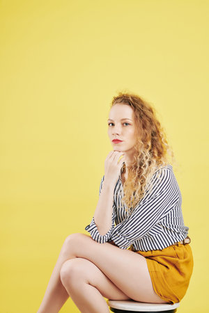 Portrait of serious young Caucasian woman with bright red lips sitting on stool and looking at camera, isolated on yellowの写真素材