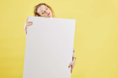 Sleepy tired young Caucasian woman leaning on big white empty paper placardの写真素材