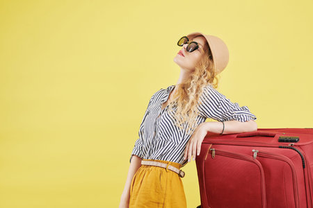 Pretty young woman in summer clothes, hat and sunglasses leaning on big red suitcase, isolated on yellowの写真素材