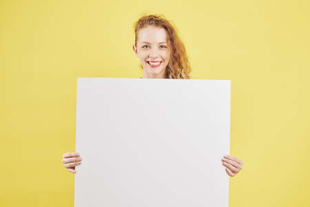 Happy pretty young curly woman holding white empty paper placardの写真素材