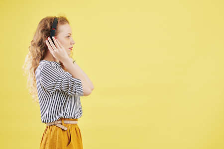 Pretty young woman in headphones standing against yellow background, listening to audio book and looking in front of herの写真素材