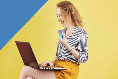 Excited young woman sitting with computer on laps and shopping with credit card for online purchasesの写真素材