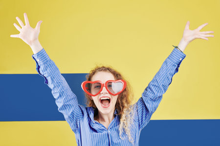 Pretty happy girl in heart shaped glasses rising arms and shouting at camera, isolated on blue and yellowの写真素材