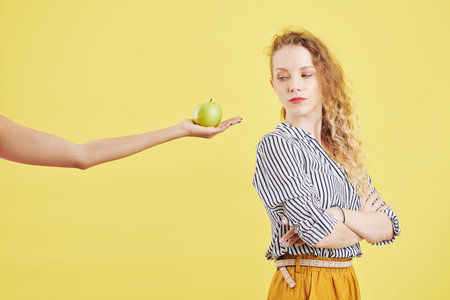 Resentful young woman folding arms and looking back at outstreched hand with green appleの写真素材