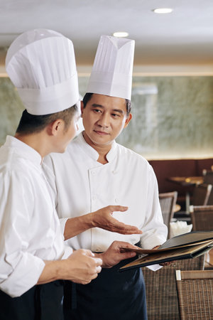 Asian chef in uniform pointing at menu and discussing it together with his colleague at the...