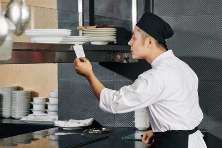 Young cook in uniform standing and reading an order on adhesive note and he is going to cook the...