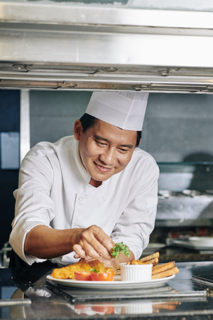 Smiling Asian mature chef prepared his brand dish and decorating it with greens in the kitchen of...