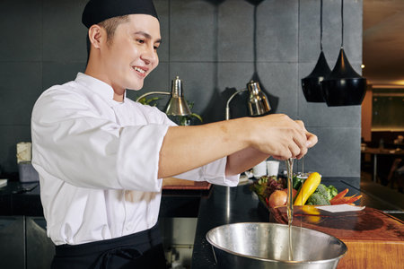Asian young cook standing and smiling while breaking eggs in the bowl in the kitchen of the...